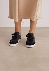 Sneaker nera con tomaia in pelle scamosciata e pelle liscia, suola intermedia bianca, lacci neri piatti e design della punta arrotondata. Indossata con una gonna di raso beige.