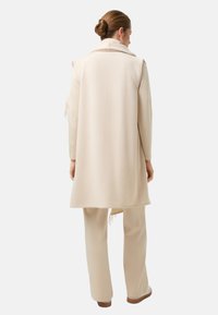 Béžová dlouhá vesta z měkké tkaniny, s límcem, detaily s třásněmi a rovný střih, doplněná o odpovídající béžové kalhoty.