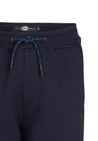 Marineblaue Jogginghose mit Kordelzugverschluss, ausgestattet mit einem strukturierten Bund und einem braunen Logo-Patch an der Seite. Glatte Stoffoberfläche.
