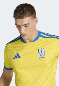 Maglia sportiva gialla con accenti blu, caratterizzata da una texture a motivi, scollo rotondo e stemma dell'Ucraina sul petto a sinistra.