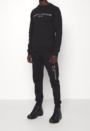 Mężczyzna ubrany w czarną bluzę Tommy Hilfiger i pasujące do niej spodnie dresowe z logo, w połączeniu z czarnymi butami na sznurowadła, stojący na tle białej ściany.