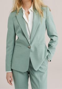 Traje a medida verde menta con cierre de un solo botón, solapas con muesca y bolsillos delanteros, combinado con una camisa blanca con cuello debajo.