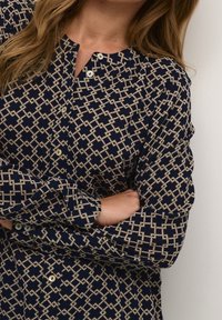 Chemise boutonnée bleu marine arborant un motif géométrique crème, à manches longues et col avec des boutons blancs. Tissu doux avec une finition texturée.