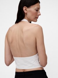 Balta petnešėlių palaidinė su aptariu, trumpu dizainu, lygiu satino paviršiumi, atvira nugara ir minimaliais siūlėmis; derinama su juodomis kelnėmis.