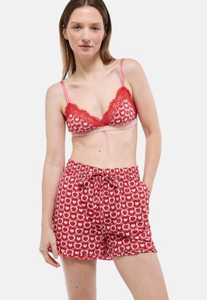 Femme portant un ensemble de lingerie rouge et rose à motifs de cœurs avec des bordures en dentelle, posant devant un fond clair uni.