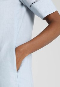 Ljusblå linnetopp med korta ärmar, med sidofickor och detaljerad sömnad vid ärmkanterna. Slät struktur.