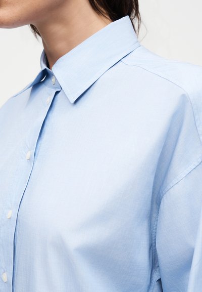 Γυναίκα με ανοιχτό γαλάζιο πουκάμισο με κουμπιά, φαρδιά γιακά και λεία υφή υφάσματος, εμφανίζεται από τους ώμους έως τον λαιμό.