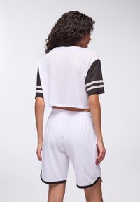 Top corto bianco in rete con maniche nere, caratterizzato da due righe bianche. Abbinato a pantaloni corti bianchi in rete, entrambi progettati per l'abbigliamento sportivo.