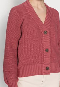 Sweter kardigan w kolorze przygaszonego różu z głębokim dekoltem w kształcie litery V, ściągaczami na mankietach i dole, z trzema brązowymi guzikami oraz teksturowaną tkaniną.