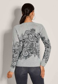 Eine Frau mit dunklem Haar im Pferdeschwanz trägt einen hellgrauen Pullover mit komplizierten schwarzen Blumen- und Paisleymustern auf dem Rücken und den Ärmeln, kombiniert mit dunklen Jeans.
