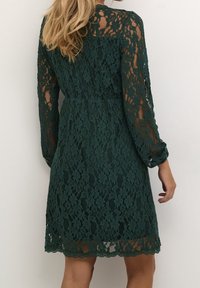 Femme aux longs cheveux blonds portant une robe en dentelle florale vert foncé à manches longues, vue de dos sur un fond blanc.