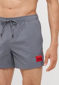 Shorts de bain gris avec taille élastique et cordon de serrage, arborant un patch rectangulaire rouge avec le logo "HUGO". Texture légère et lisse.