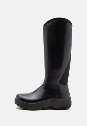 Bota alta de goma negra con puntera redondeada y suela gruesa, acabado liso y un panel elástico en la parte superior trasera para un ajuste ceñido.
