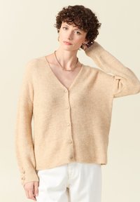 Cardigan beige à côtes, décolleté en V, fermeture à boutons, manches longues avec accents en boutons. Texture douce, coupe décontractée, design superposé.