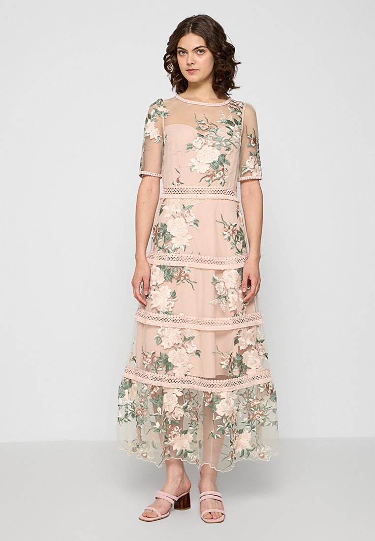 Abito lungo di un morbido rosa con sovrapposizione floreale trasparente, maniche a metà lunghezza e dettagli in pizzo. Presenta un design a strati e motivi floreali ricamati.