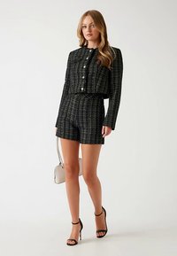 Ensemble coordonné en tweed noir comprenant une veste courte à boutons et un short assorti. Comprend un sac à main structuré et des talons à brides.