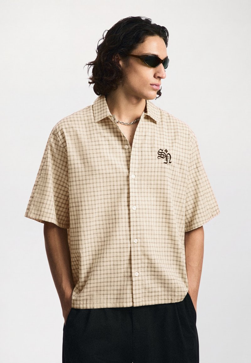 Jeune homme aux cheveux bouclés foncés porte des lunettes de soleil marron, une chemise beige à carreaux à manches courtes avec un petit logo noir et un pantalon noir.