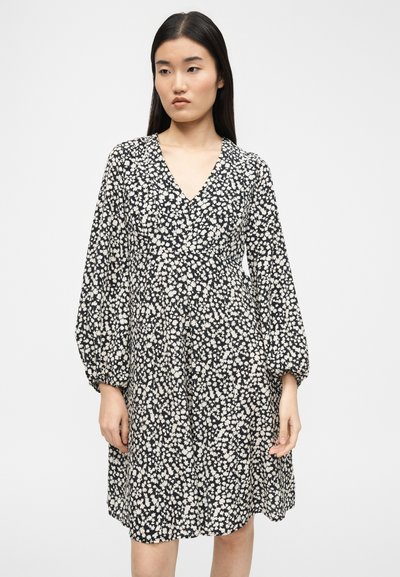Νεαρή γυναίκα που φοράει μαύρο φόρεμα με λευκό ανθισμένο σχέδιο, μακριά μανίκια μπαλόνι και V-λαιμόκοψη, στέκεται μπροστά σε απλό λευκό φόντο.