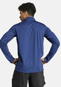 Marineblauw sportshirt met lange mouwen, rechte kraag, gladde textuur en subtiele stiksels, gedragen met de handen op de heupen.