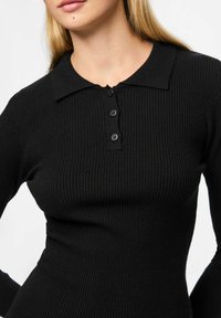 Žena nosi crnu rebrastu pletivu dugom rukavu polo majicu s tri crne dugmadi, prikazana od vrata do struka na bijeloj pozadini.