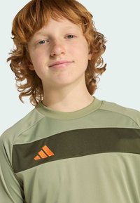 Olivově zelené sportovní tričko s tmavě zeleným vodorovným pruhem, s oranžovým třípruhým logem Adidas na levé straně. Hladký povrch.