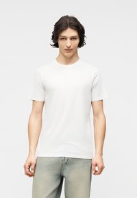 Νεαρός άνδρας με σκούρα, κυματιστά μαλλιά φορώντας ένα απλό λευκό κοντομάνικο μπλουζάκι και ανοιχτόχρωμα μπλε τζιν, που στέκεται μπροστά σε ουδέτερο φόντο.