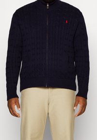 Marinblå stickad tröja med hög krage, som har dragkedjeslutning, texturerade kabelmönster och ribbade ärmslut och nederkant. Röd logotyp på bröstet.