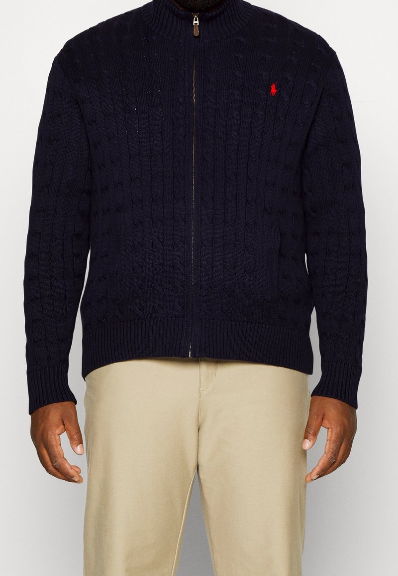 Marinblå stickad tröja med hög krage, som har dragkedjeslutning, texturerade kabelmönster och ribbade ärmslut och nederkant. Röd logotyp på bröstet.