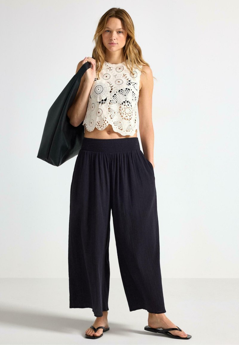 Top corto bianco all'uncinetto con motivi floreali, abbinato a pantaloni neri a gamba larga e infradito neri. Modella con una borsa scura.