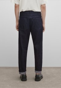 Tummansiniset suoralahkeiset farkut, näkyvät takataskut, käännetyt lahkeet ja sileä rakenne. Päällä mustat kengät.