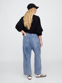 Blonde sieviete melnā cepurē, melnā sporta krekliņā, brīvās zilajās džinsās un baltajās kedās stāv ar muguru izgriezusies, ar vienu roku aizmugurējā kabatā.