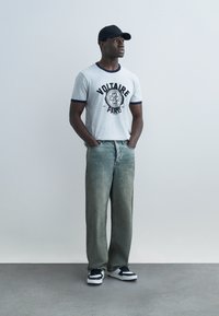 Man draagt een grijs Voltaire Paris T-shirt, wijde, vervaagde jeans, zwart-witte sneakers en een zwarte baseballpet, staand met handen in de zakken.