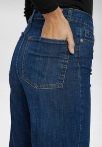 Dark blue denim jeans with a fitted silhouette, featuring a visible back pocket and contrasting yellow stitching details.