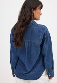 Camisa de denim oversized en azul oscuro, con botones en la parte delantera, mangas largas y dobladillo curvado. Presenta un yugo y un detalle de pliegue en la parte trasera.