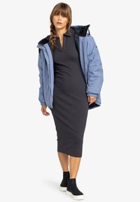 Doudoune bleue avec capuche, portée par-dessus une robe gris foncé. La doudoune présente des poignets élastiques et une texture brillante ; associée à des bottes noires à plateforme.