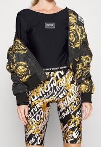 Svart bodysuit med en Versace Jeans Couture-logotyp, tillsammans med en bomberjacka med gyllene barockmönster och svarta åtsittande shorts med vit text.