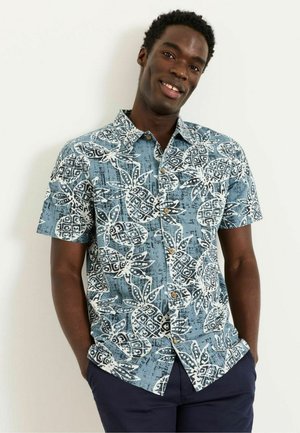 Mann trägt ein blaues Kurzarm-Hemd mit weißen und schwarzen Ananasmustern, steht mit den Händen in den Taschen und lächelt.