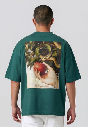 Persoon die een groen shirt draagt met een afbeelding van een hand die een appel vasthoudt en een slang op bladeren met de tekst "De Oerzonde" op de achterkant.