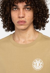 Beige bomuld t-shirt med rund halsudskæring og et cirkulært logo med teksten "Jorden, Vinden, Ilden, Vandet" i hvidt på venstre brystområde.