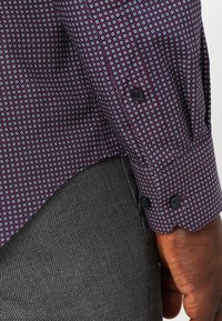 Bordeauxfarbenes Hemd mit geometrischem Muster, langen Ärmeln mit schwarzen Knöpfen und glatter Textur, kombiniert mit grauem Stoff.