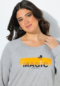 Donna con capelli scuri che indossa orecchini a cerchio argentati e una maglietta grigia con la scritta "MAGIC FEELING" su una pennellata gialla.