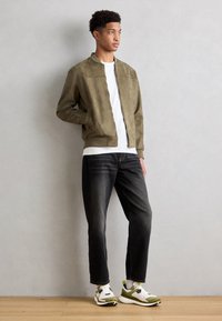 Olivgrüne Bomberjacke mit Reißverschluss auf der Vorderseite, kombiniert mit schwarzen Jeans und weißen Sneakers, die mit grünen und schwarzen Details akzentuiert sind.