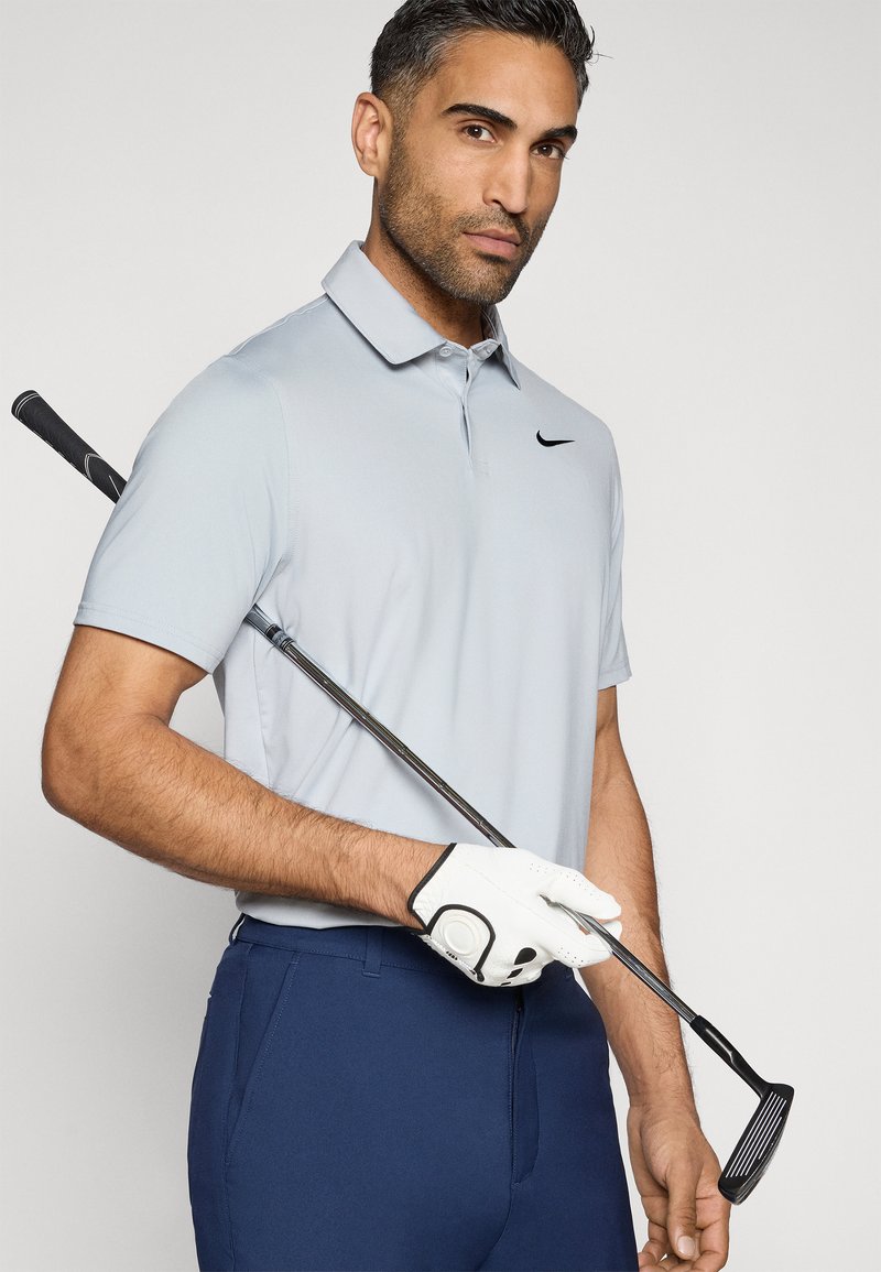 Hellblaues Poloshirt mit kurzen Ärmeln, schwarzem Logo, dunkelblauen Hosen, weißem Golfhandschuh, der einen Golfschläger mit schwarzem Griff hält.