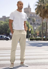 Beige stretchbroeken met elastische boorden, gecombineerd met een aansluitend wit T-shirt en lichtgrijze instapschoenen, tegen een stedelijke achtergrond.