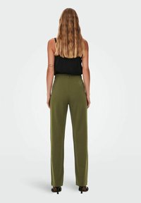 Žena s dlouhými vlnitými blond vlasy má na sobě černý bezrukávový top, vysoké olivové široké kalhoty a černé podpatky, stojí čelem k odvrácené straně.