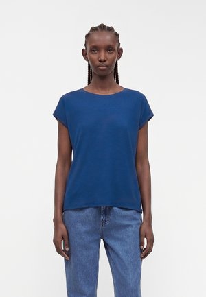 Kvinde med fletninger står vendt fremad iført en ensfarvet blå kortærmet top og lyseblå denimjeans mod en hvid baggrund.