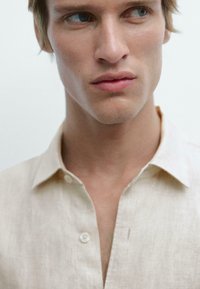 Light beige linen shirt with a collar, subtle texture, and a front button closure. Visible stitching detail at the neckline.