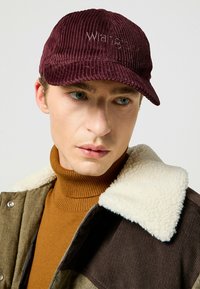 Mörkröd corduroykeps med synlig ribbing, prydd med en broderad "Wrangler"-logotyp. Modellen bär en brun polo och en jacka med fårullsdetalj.