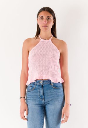 Giovane donna con capelli castani lisci che indossa un top halter rosa lavorato a maglia e jeans blu, in piedi davanti a uno sfondo bianco semplice.