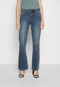 Žena v bledě modrých bootcut džínách a černých páskových lodičkách na vysokém podpatku, stojící na neutrálním světlém pozadí.